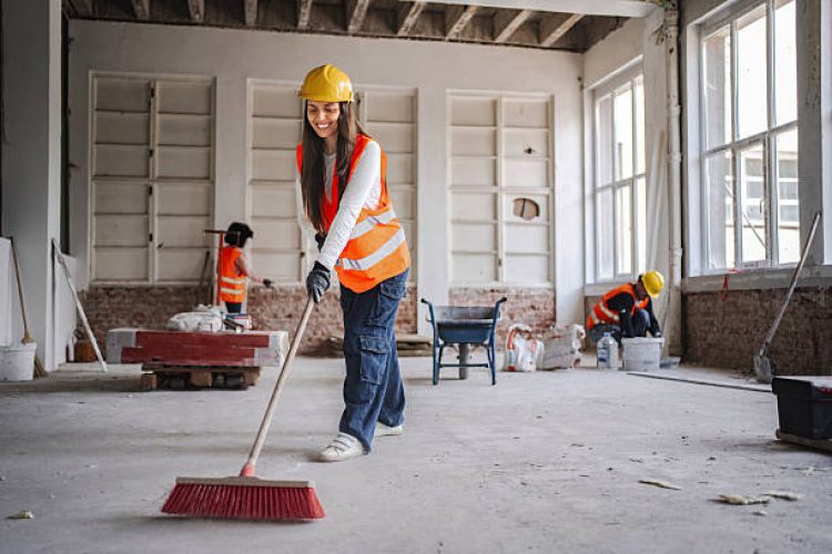 Smiling female construction worker sweeping the floor on a construction site while her colleagues are working