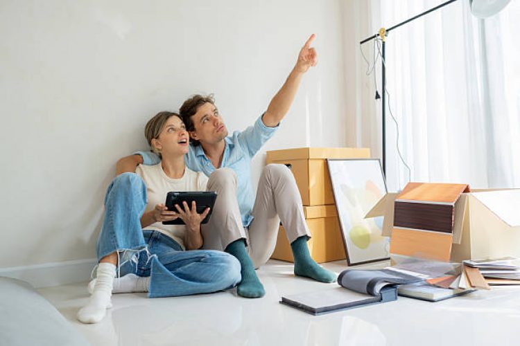 Amid a messy room full of moving boxes, a couple relaxes on the floor with a tablet, excitedly talking about decorating their new space. A cozy moment of bonding, teamwork, and creative planning for their new life together.