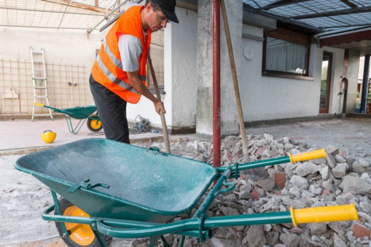 e12 Home repair. Removal of debris, concrete blocks and bricks from the bottom of a roof - terrace. Worker with orange reflective jacket and shovel in his hand