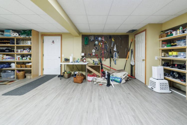 Spacious basement features shelving units filled with household items. Central area includes pegboard with hanging tools, laundry baskets, and home maintenance supplies. Light wood-look flooring and drop ceiling enhance the clean, functional space. Doors lead to additional storage or utility areas. Space serves multiple purposes for storage, laundry, and home improvement projects.