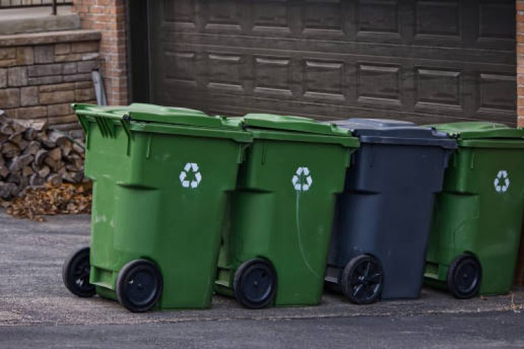 Four bins are aligned against a closed garage wall. Three are green recycling bins with recycling symbols, and one is black, likely for waste. Each bin has two wheels for easy movement.